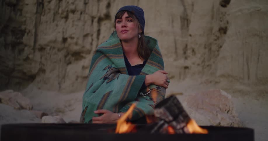 A thoughtful woman wrapped in a patterned blanket sits alone by a campfire in a remote desert canyon