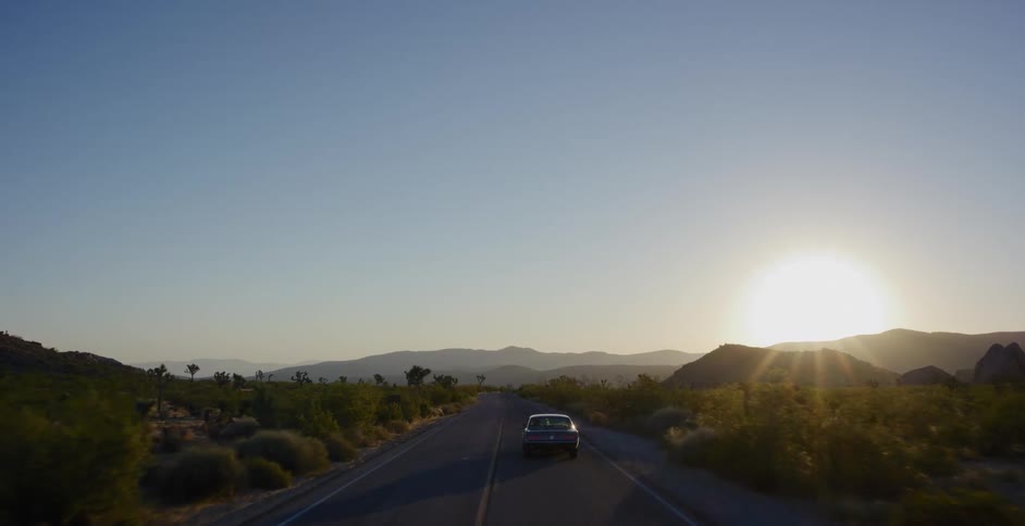 A vintage car drives along a scenic desert road in Joshua Tree National Park towards the golden setting sun