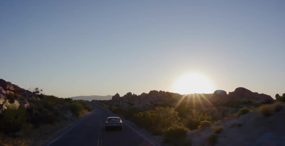 An aerial shot follows a classic car as it drives along a scenic desert highway into the golden sunrise