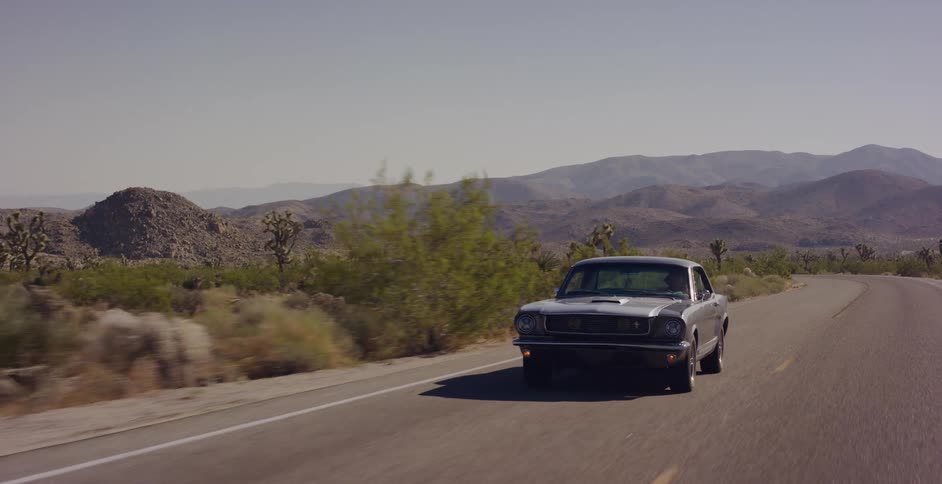 A classic silver Ford Mustang drives down a winding desert highway with mountains in the background