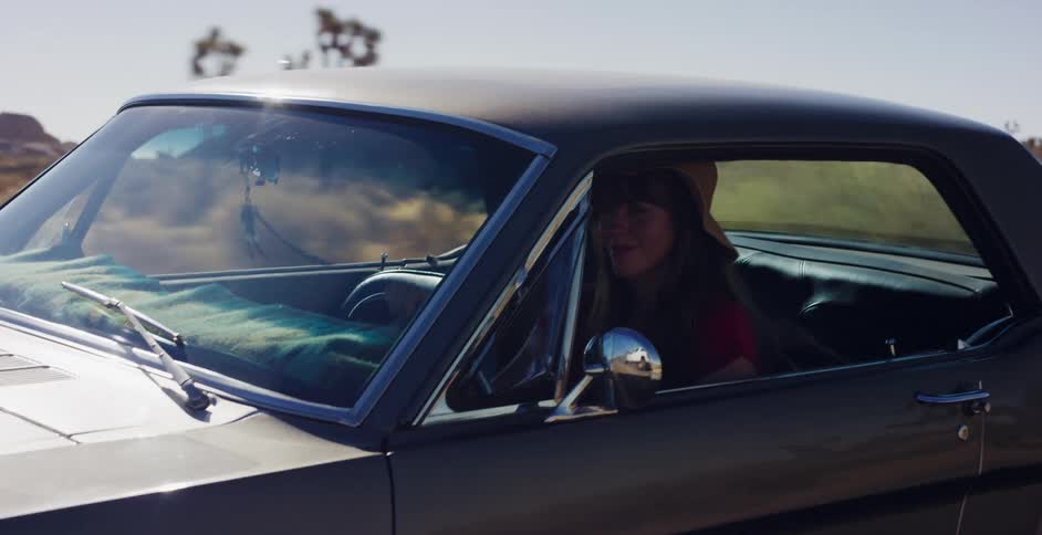 A woman in a sun hat drives her classic car down a dusty desert road in slow motion