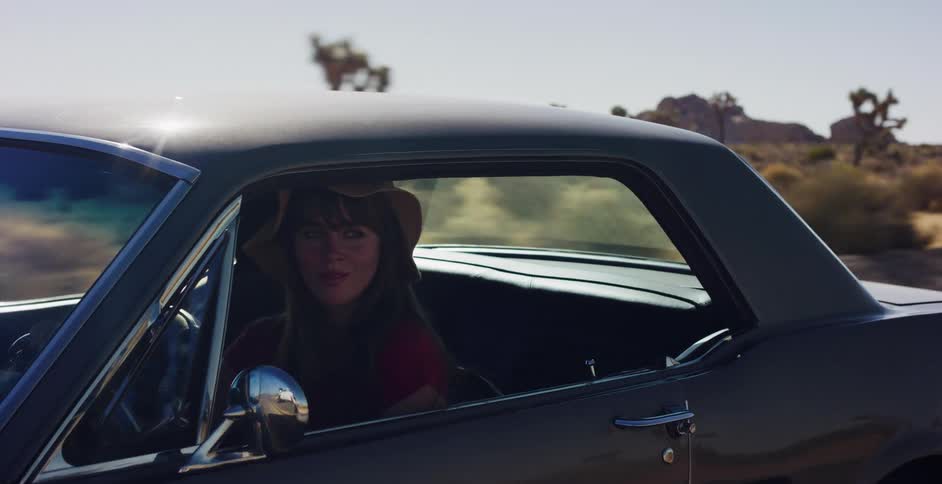 A young woman in a hat smiles while driving a classic Mustang down a dusty desert road in slow motion