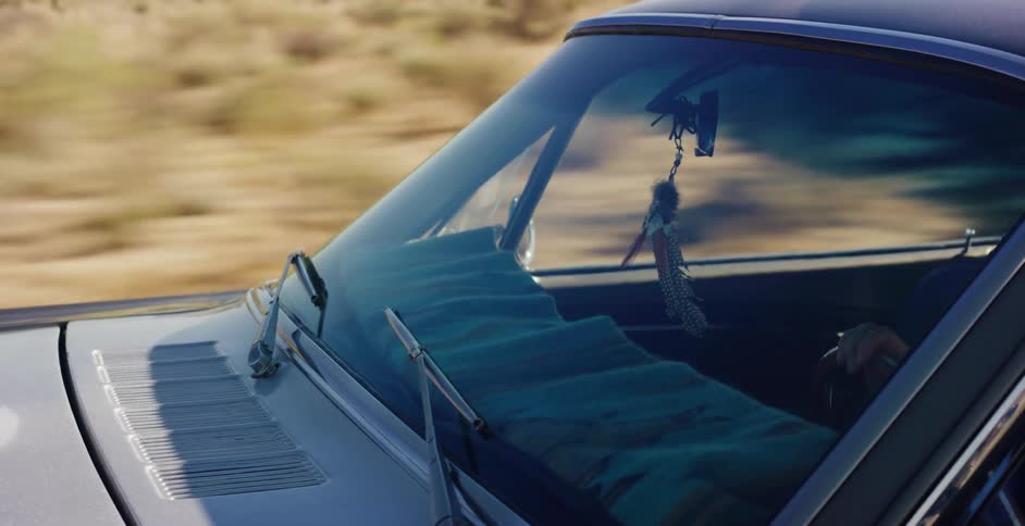A dreamcatcher hangs from the rearview mirror of a classic car speeding down a desert road