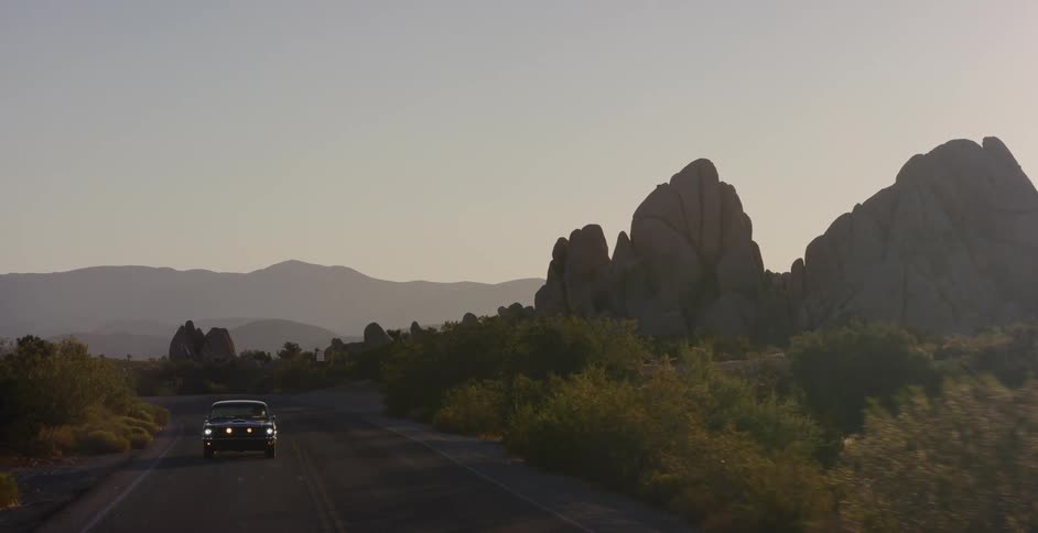 A classic black car cruises down a winding desert road through rocky landscapes at sunrise