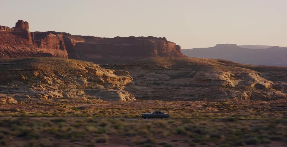 A classic American muscle car drives on a scenic desert highway through a majestic canyon landscape at golden hour