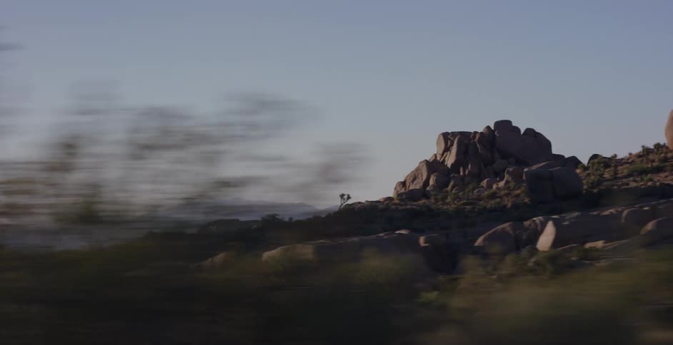 A scenic driving shot through a desert landscape with large rock formations and mountains in the distance at golden hour