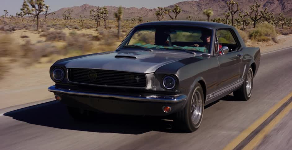 A classic grey Ford Mustang cruises down a desert highway surrounded by Joshua trees and mountains on a sunny day