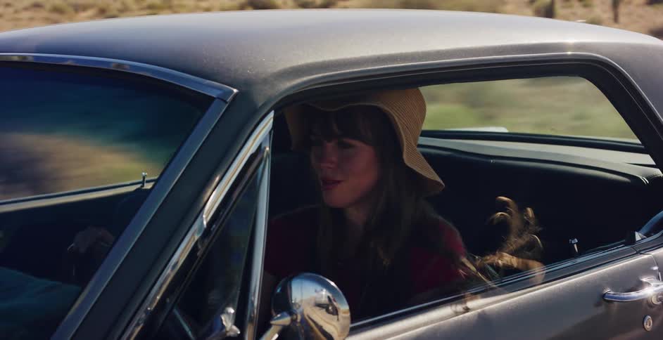 A young bohemian woman with long hair drives a classic silver car down a desert highway on a sunny day
