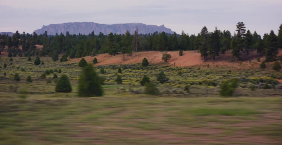 A scenic driving shot of a rugged, tree-covered landscape with distant mountains under a cloudy sky