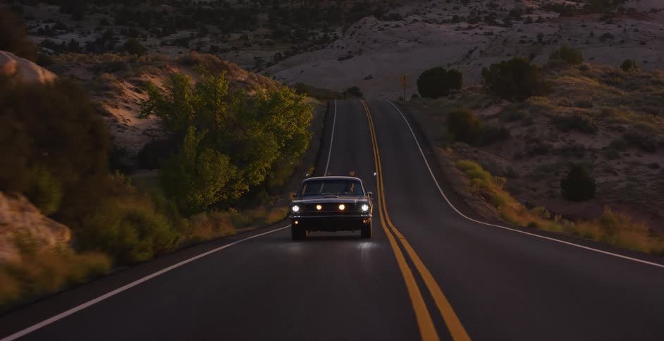 A classic black Ford Mustang with its headlights on drives down a remote desert highway at dusk