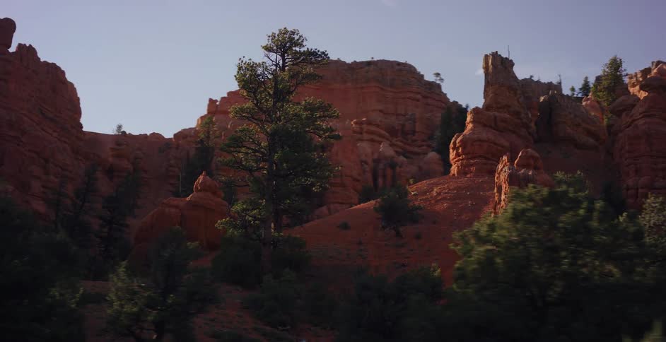 A slow-motion driving shot reveals stunning red rock canyon formations dotted with evergreen trees, ending with a cinematic lens flare