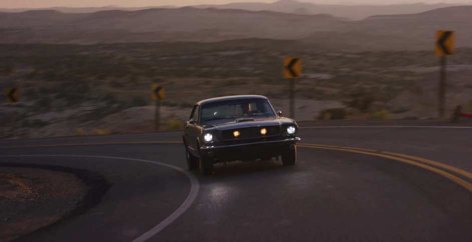 A vintage black Ford Mustang drives along a winding desert highway at sunrise