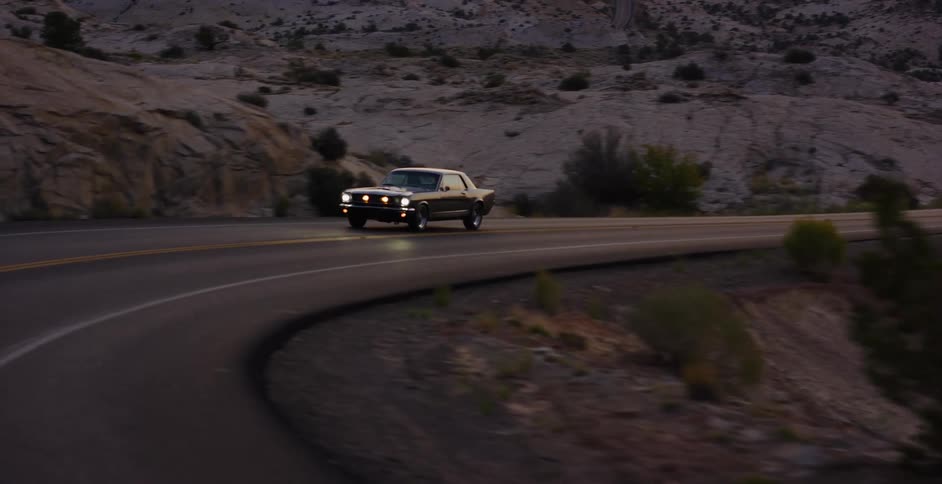 An aerial view tracks a vintage black sports car as it navigates a winding desert highway at dusk