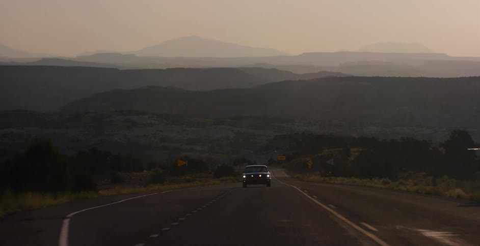Drone pulls back revealing a car driving along a winding desert road with hazy mountains in the background