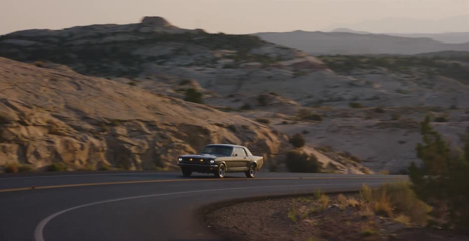 A classic silver Ford Mustang cruises along a winding desert road through a rocky landscape at golden hour