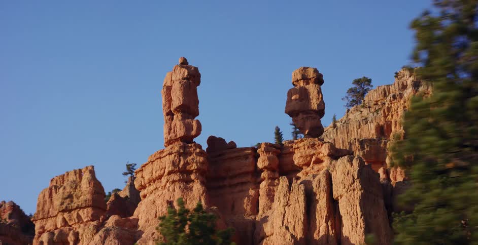 Tracking shot moving past two unique red rock hoodoo formations against a clear blue sky in a canyon
