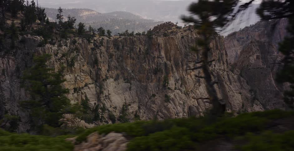 A smooth dolly shot reveals a vast, rocky mountain landscape with pine trees and shrubs in the foreground