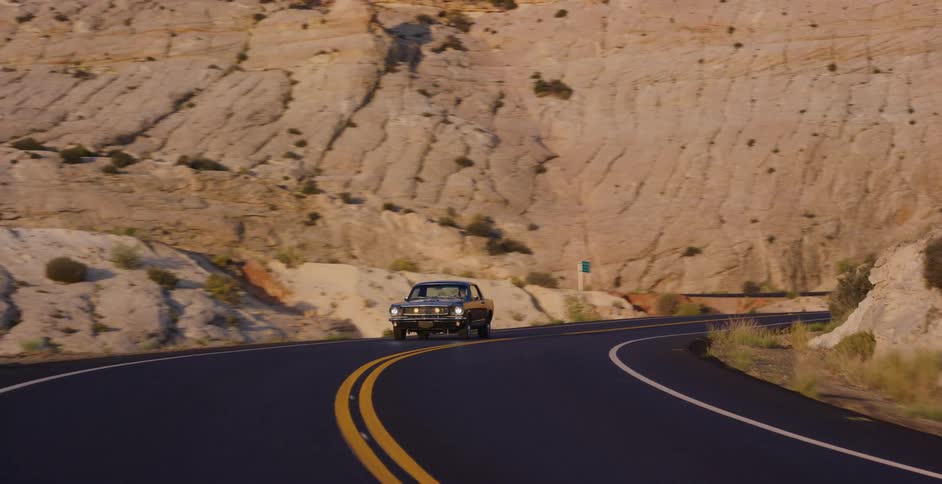 A classic muscle car cruises down a scenic, winding road through a rocky desert canyon during golden hour