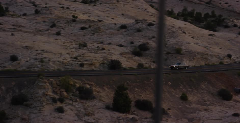 A vintage car with its headlights on navigates a winding road through a stark desert canyon landscape at twilight