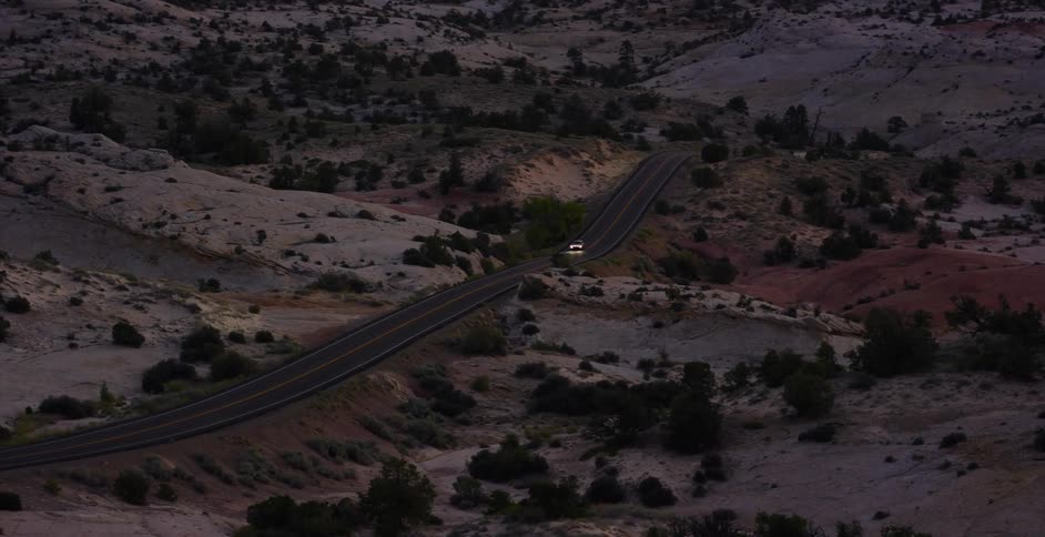 A car drives down a winding road through a rocky desert canyon at dusk with its headlights on