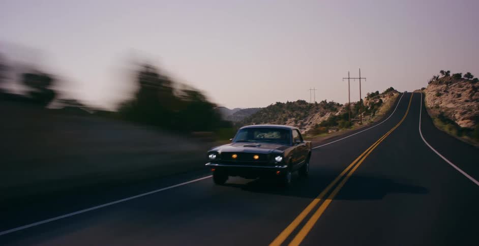 A vintage black muscle car cruises along a scenic and winding mountain road during a beautiful sunset