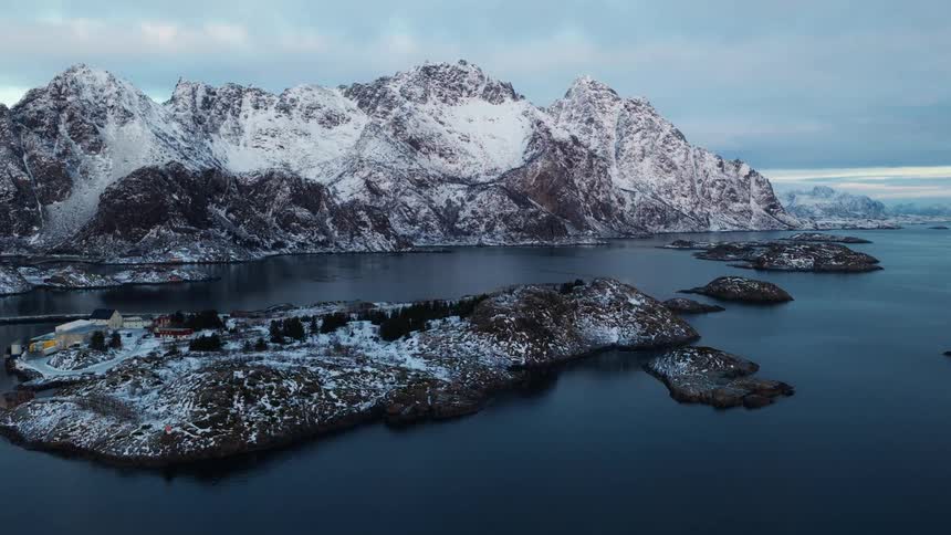 An epic aerial view flies towards a small snow-covered village on a remote island surrounded by tranquil waters and majestic mountains in Norway