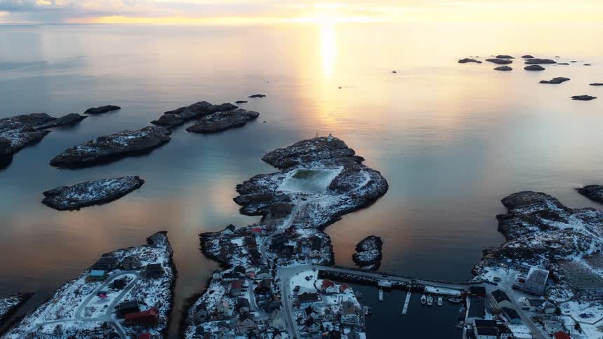 Aerial pullback shot of a remote, snow-covered island village and its unique football pitch surrounded by the tranquil ocean at sunset