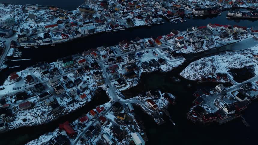 A slow aerial flyover reveals a tranquil, snow-covered coastal town nestled among dark waters at dusk