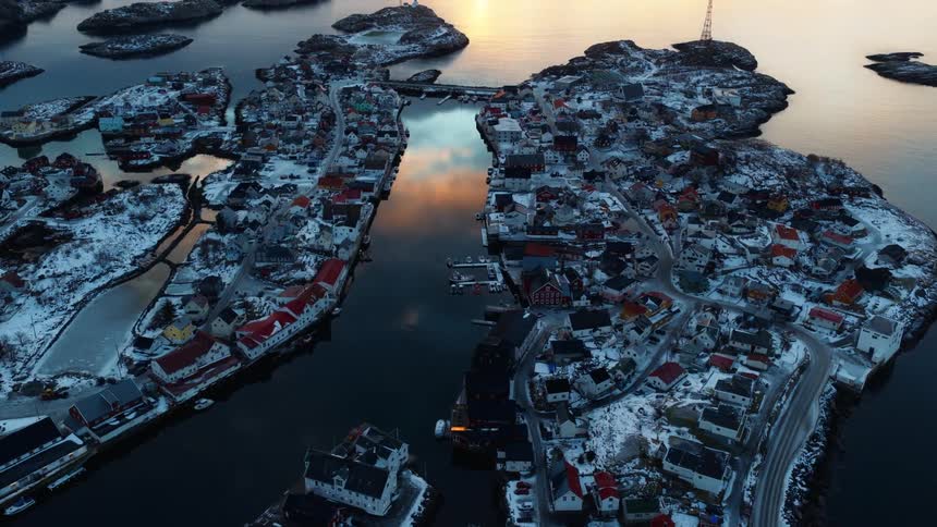 A serene aerial pull-back reveals a picturesque, snow-covered fishing village on a cluster of islands during a golden sunrise in Norway