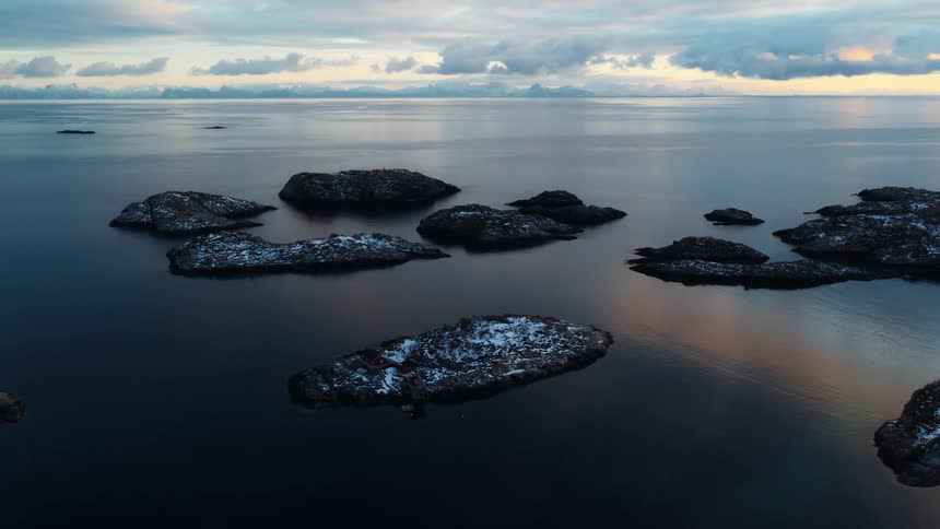 A stunning aerial view shows a tranquil archipelago of small, snow-dusted islands in a calm sea at sunset