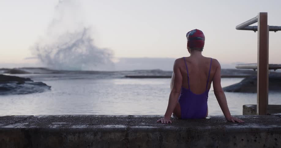 A female swimmer sits contemplatively by a tidal pool as a large ocean wave crashes dramatically in slow motion