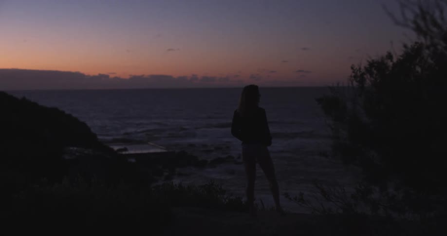 A silhouetted woman peacefully watches the ocean from a coastal cliff during a serene twilight sunset