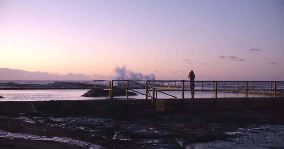A woman in a swimsuit watches the ocean waves from a pier beside a tranquil rock pool during a beautiful sunrise