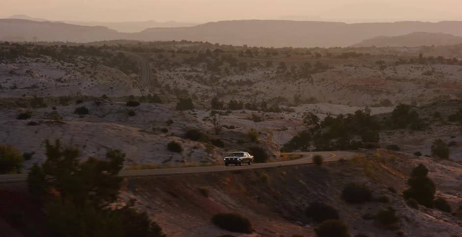 A classic muscle car cruises down a winding desert road at sunrise, revealing a beautiful scenic landscape