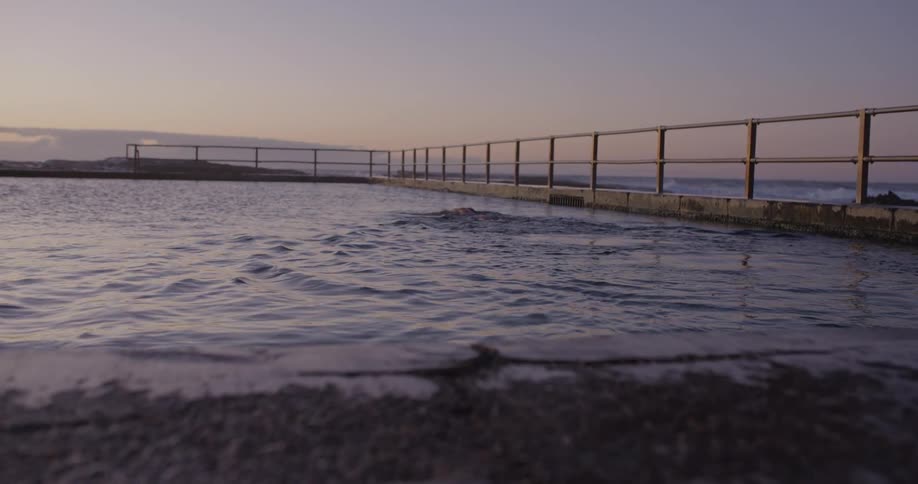 A woman swims freestyle laps in slow motion in a tranquil ocean pool as the sun rises, with waves crashing in the background