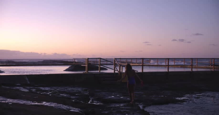 In slow motion, a woman in a one-piece swimsuit walks away from the camera across a rocky shoreline toward an ocean pool during a pastel sunrise
