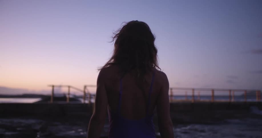 A woman in a purple swimsuit is silhouetted against the dusky sky as she stands on a rocky beach, her hair blowing in the wind in slow motion