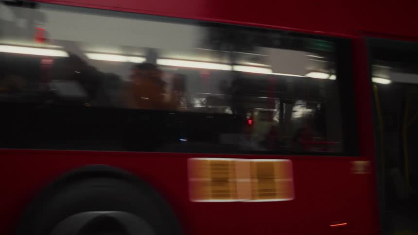 A modern red double-decker city bus drives past the camera through a tree-lined intersection at dusk