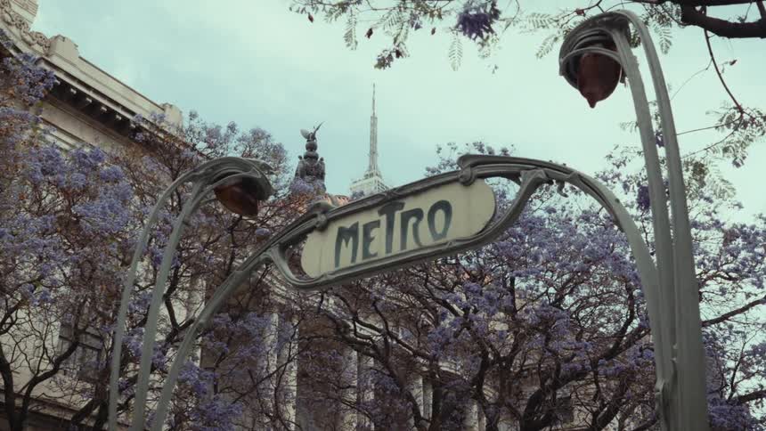 An Art Nouveau Metro sign stands before the Palacio de Bellas Artes in Mexico City, surrounded by blooming purple jacaranda trees