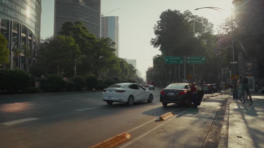 Slow-moving traffic flows down a sunlit boulevard in Mexico City as pedestrians and cyclists go about their day