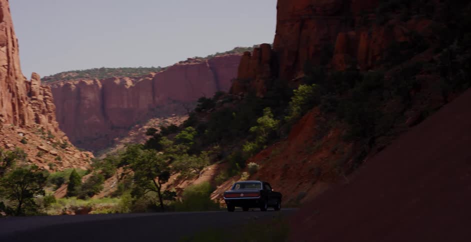 A classic dark muscle car slowly drives away on a winding road through a dramatic red rock canyon