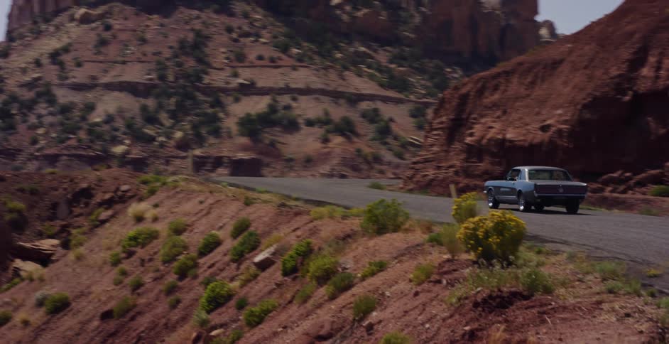 A classic muscle car cruises down a winding desert road through a stunning red rock canyon