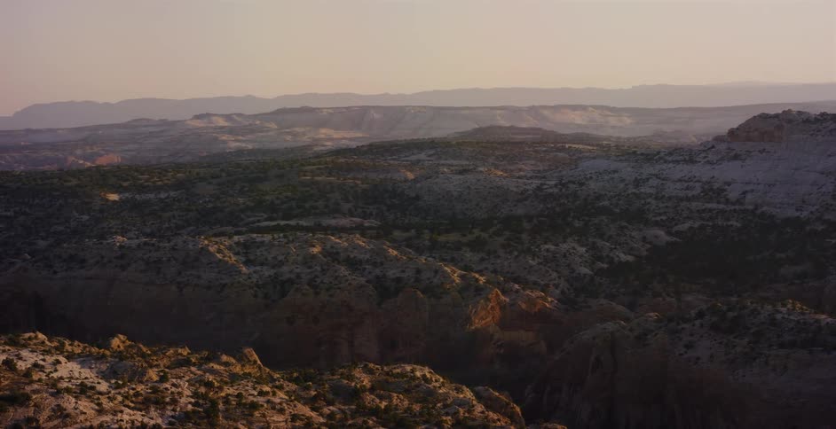 A scenic view from a moving vehicle captures the vast, rocky desert canyon landscape during a hazy golden hour