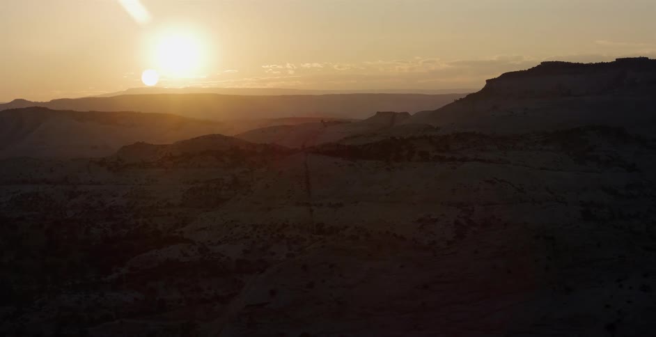 A slow aerial shot flies over a vast desert canyon landscape as the sun sets, casting a golden glow