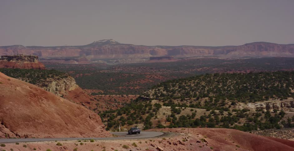A pickup truck drives along a winding road through a vast and scenic red rock canyon landscape