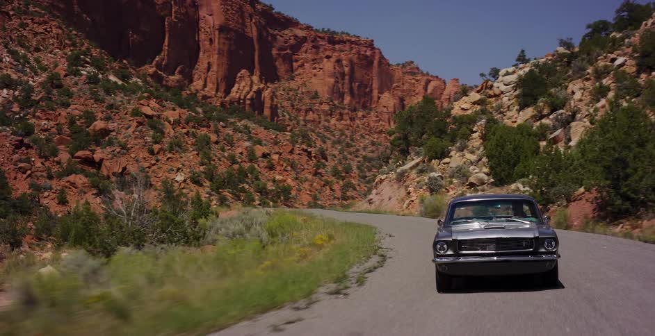 A classic silver Mustang cruises down a winding desert road surrounded by majestic red rock canyons