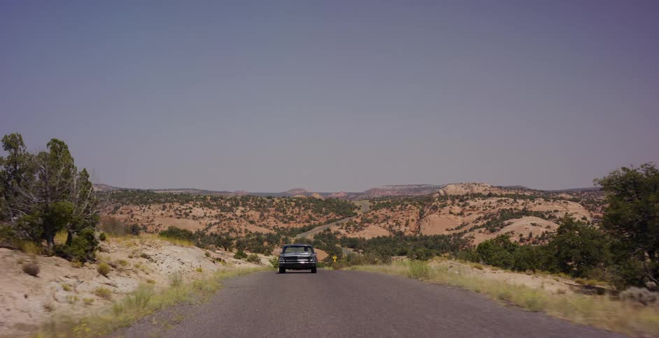 A vintage car slowly drives towards the camera on a remote desert highway in the American Southwest