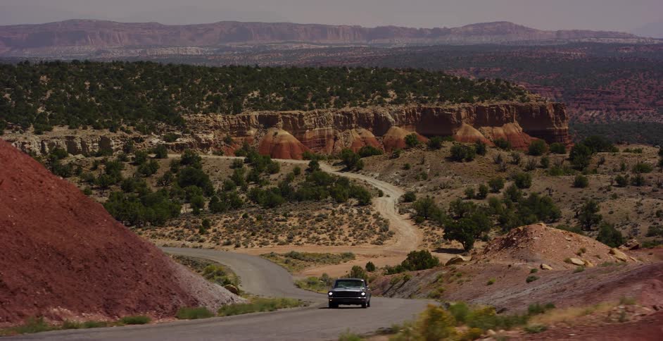 A classic black Ford Mustang drives along a winding road through the scenic red rock desert landscape of Utah