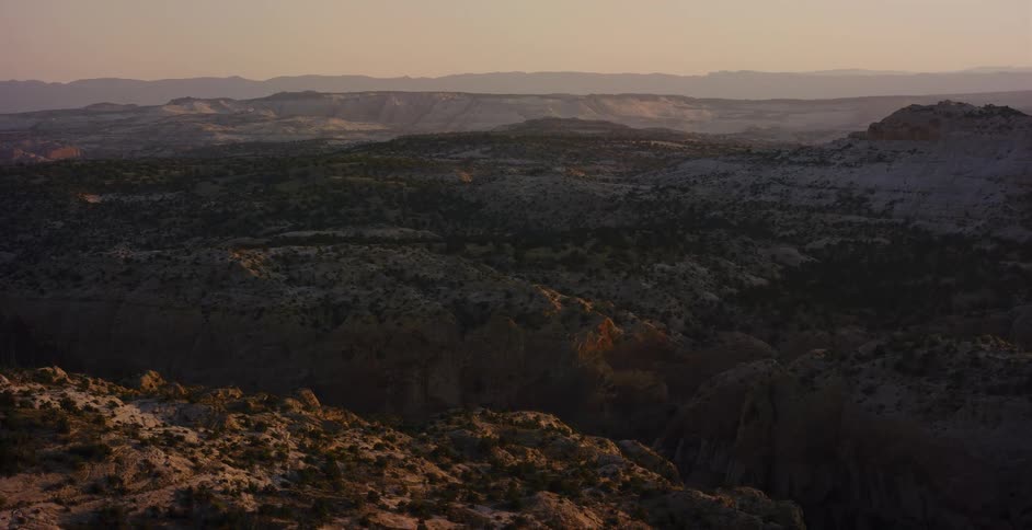 An expansive aerial view pulls back over a rugged desert canyon landscape bathed in the warm light of a golden hour sunset