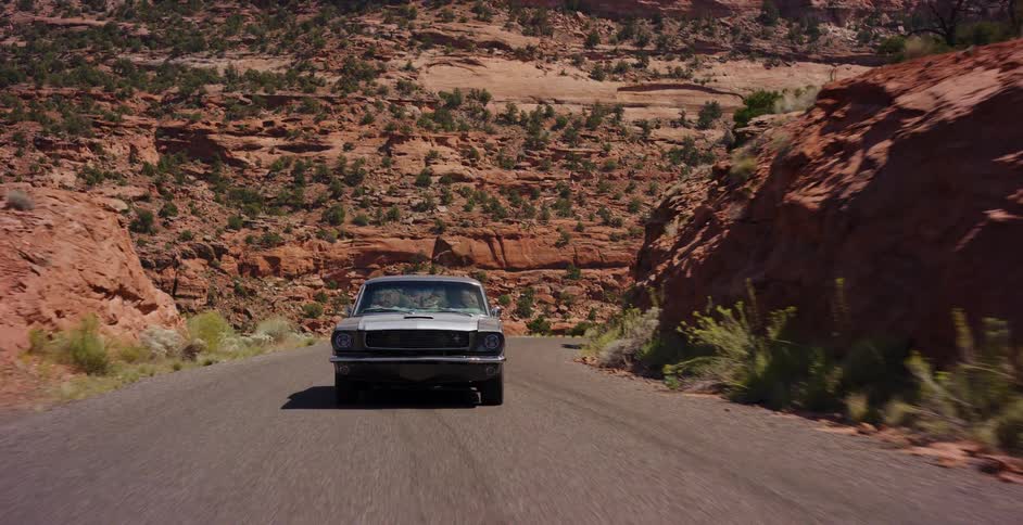 A vintage silver muscle car drives along a winding road through a stunning red rock desert canyon